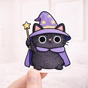 May include: Embroidered patch of a black cat in a wizard costume. The cat has a purple wizard hat with yellow stars and a matching cape. It holds a gold star-tipped wand. The patch has a white border.
