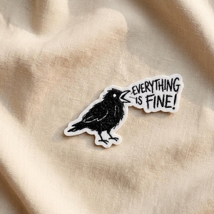 May include: A black crow patch with the text "EVERYTHING IS FINE!" in a speech bubble. The crow is facing left with its beak open. The patch is on a beige fabric background.