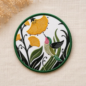 May include: A circular embroidered patch featuring a white background with a green border. The design depicts a hummingbird with a pink throat and green body approaching yellow fan-shaped flowers surrounded by dark green foliage and stems.