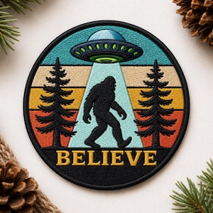 May include: Embroidered patch featuring a black Bigfoot silhouette being beamed up by a teal UFO. The design includes a retro sunset background with the word "BELIEVE" in gold lettering. Two pine trees frame the scene.