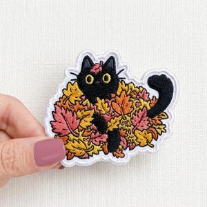 May include: Embroidered patch featuring a black cat nestled in colorful autumn leaves. The cat has yellow eyes and a black tail. The leaves are shades of orange, yellow, and pink, with a white border.