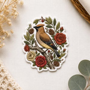 May include: Embroidered patch featuring a bird perched on a branch, surrounded by red and yellow roses, green leaves, and pinecones. The bird has brown, tan, and black feathers. The patch has a white scalloped border, perfect for adding to clothing or accessories.