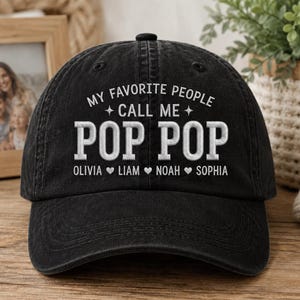 May include: Black baseball cap with the text "MY FAVORITE PEOPLE CALL ME POP POP" in white. Below the text are the names Olivia, Liam, Noah, and Sophia. The cap has a curved brim and a button on top.