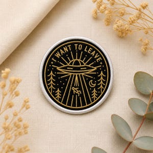 May include: Round black and gold embroidered patch with the text "I WANT TO LEAVE". The design features a UFO, trees, a person being beamed up, and a crescent moon. The patch has a white border.