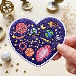 May include: A heart-shaped embroidered patch with a dark purple background featuring space-themed motifs including a ringed planet, a UFO with an alien, Earth, a satellite, a galaxy, a comet, a crescent moon, and yellow stars.