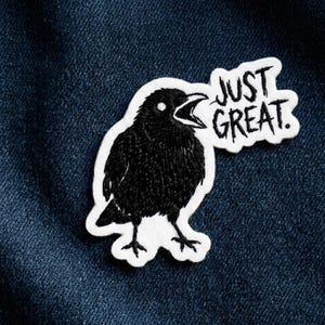 May include: Embroidered patch featuring a black crow with an open beak, accompanied by the words "JUST GREAT." The patch has a white border and is set against a dark blue denim background. The crow is detailed with textured stitching.