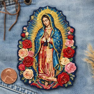 May include: Embroidered patch depicting a religious figure in a blue robe with a gold halo, encircled by red and pink roses. The patch is on denim, with a penny for scale.