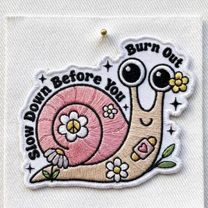 May include: Embroidered patch featuring a cartoon snail with large eyes and a smiling face. The snail's shell is pink with a white daisy in the center. Text reads "Slow Down Before You Burn Out".