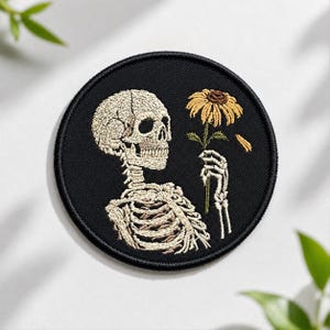 May include: A circular embroidered patch featuring a beige skeleton on a black background. The skeleton is holding a yellow sunflower with a green stem. The patch has a black stitched border and a detailed design of the skull and rib cage.
