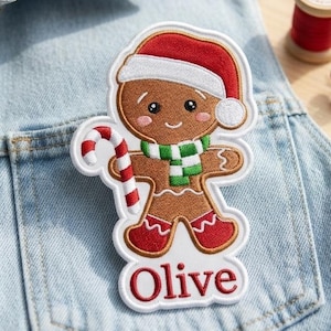 May include: An embroidered patch featuring a brown gingerbread person wearing a red and white Santa hat, a green and white striped scarf, and red boots. The figure holds a red and white candy cane, with the name Olive stitched in red below.