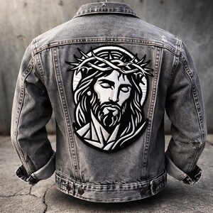 May include: A gray denim jacket with a large, embroidered patch on the back. The patch features a black and white illustration of Jesus wearing a crown of thorns. The jacket has a classic design with button closures and pockets.