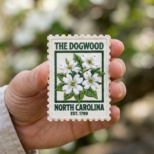 May include: Embroidered patch featuring a detailed depiction of dogwood flowers. The patch reads "THE DOGWOOD" at the top and "NORTH CAROLINA EST. 1789" at the bottom, with a green border and white flowers. The patch is held in a hand.
