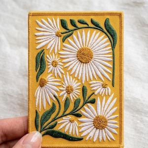 May include: Embroidered patch with a daisy floral design. The patch has a yellow background with white daisies and green leaves. The daisies have yellow centres. The patch is rectangular with a stitched border.