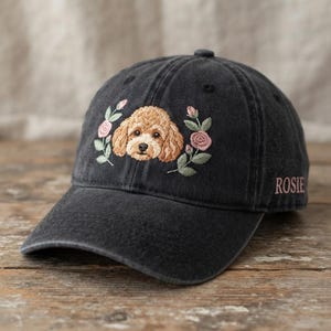 May include: A charcoal black baseball cap featuring an embroidered tan poodle face surrounded by pink roses and green leaves. The name ROSIE is embroidered in pink thread on the side of the cap. The cap has a curved brim and a structured crown.