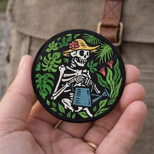 May include: Round black patch featuring a skeleton wearing a straw hat and watering plants with a blue watering can. The skeleton is surrounded by green leaves and red flowers. The patch has a black border.