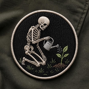May include: Embroidered patch featuring a skeleton kneeling and watering a small plant with a watering can. The patch is circular with a black background and a cream-colored border. The skeleton and plant are detailed.