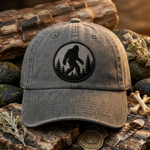 May include: A charcoal gray baseball cap featuring a circular embroidered patch on the front. The patch displays a black silhouette of a Bigfoot creature walking through a forest of pine trees against a light gray background.