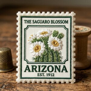 May include: Embroidered patch in the shape of a postage stamp featuring the text "THE SAGUARO BLOSSOM" and "ARIZONA EST. 1912". The patch depicts white flowers with yellow centers and green cactus. The border is green.
