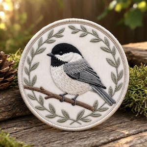 May include: Embroidered patch featuring a chickadee bird perched on a branch, surrounded by a laurel wreath. The bird has black, white, and gray feathers. The patch is circular, with a cream-colored background and a brown branch. The wreath is green.