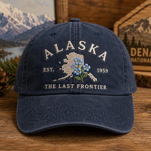 May include: Navy blue baseball cap with embroidered "ALASKA" text, a map outline, and blue floral accents. The cap also features "EST. 1959" and "THE LAST FRONTIER" text. A stylish accessory for outdoor enthusiasts.