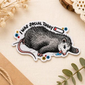 May include: Embroidered patch featuring a gray opossum with the text "I WAS SOCIAL TODAY." The opossum has its tongue out and is surrounded by colorful floral accents. The patch is white with a stitched border.