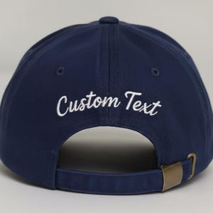 May include: Navy blue baseball cap with the embroidered text "Custom Text" in white. The cap has a curved brim, a brass-colored metal clasp, and an adjustable strap. The cap is a classic style.