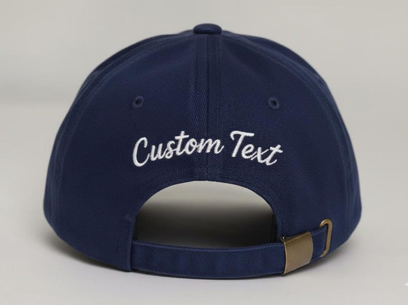 May include: Navy blue baseball cap with the embroidered text "Custom Text" in white. The cap has a curved brim, a brass-colored metal clasp, and an adjustable strap. The cap is a classic style.