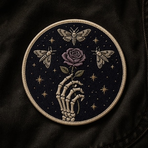 May include: Embroidered patch featuring a skeleton hand holding a purple rose. Three moths and gold stars are embroidered on a navy blue background. The patch has a beige border and is circular in shape.