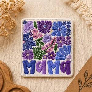 May include: Embroidered patch featuring a floral design in shades of purple and blue, with the word "MAMA" in a stylized font. The patch has a white border and is likely for clothing or accessories.