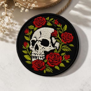 May include: A circular embroidered patch featuring a white skull centered on a black background, surrounded by a wreath of vibrant red roses and green leaves. The embroidery displays detailed stitching for the skull and floral elements.