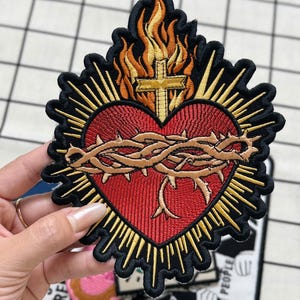 Puede incluir: Un parche bordado que muestra un corazón rojo rodeado por una corona de espinas marrón. Sobre el corazón hay una cruz amarilla rodeada de llamas naranjas y amarillas. El diseño está delineado en negro con rayos amarillos hacia afuera.