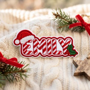 May include: A custom embroidered patch featuring the name EMILY in red and white candy cane stripes. The design includes a red Santa hat perched on the letter E and a green holly leaf with three red berries attached to the letter Y.