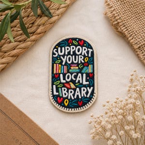 May include: Embroidered patch with the text "SUPPORT YOUR LOCAL LIBRARY" in white, yellow, and red. The patch features colorful book illustrations and decorative leaves and hearts on a navy blue background.