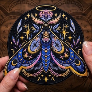 May include: Embroidered patch featuring a detailed moth design with a halo, stars, and floral elements. The moth has blue, pink, and gold accents on a black background. The patch is round and has a decorative border.