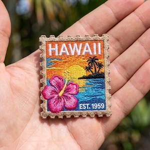 May include: Embroidered patch featuring a Hawaiian sunset scene with a pink hibiscus flower, palm trees, and the word "HAWAII". The patch is in a postage stamp design with the text "EST. 1959".
