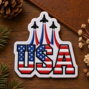 May include: A white embroidered patch featuring the letters USA in a red, white, and blue flag pattern. Above the letters, three black jet silhouettes fly in formation, each leaving behind a trail of red and blue embroidered smoke.