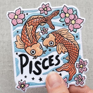 May include: Embroidered patch featuring two orange koi fish swimming in opposite directions, with the word "Pisces" in black script. The patch is bordered by pink flowers and blue water lines, with small black stars.