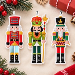 May include: Three embroidered nutcracker patches labeled 1, 2, and 3. Patch 1 is a red-coated nutcracker with blue striped pants. Patch 2 is a green-coated nutcracker holding a gold staff. Patch 3 is a green-coated nutcracker playing a red drum.