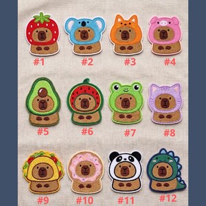 May include: A collection of twelve embroidered patches featuring a bear character in various costumes, including a strawberry, koala, fox, pig, avocado, watermelon, frog, cat, taco, donut, panda, and dinosaur. Each patch is numbered.