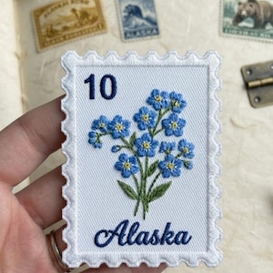 Peut inclure: Un écusson brodé rectangulaire en forme de timbre-poste avec un fond blanc et des bords festonnés. Il présente un bouquet de myosotis bleus aux centres jaunes, le chiffre 10 en bleu foncé et le mot Alaska écrit en bleu.