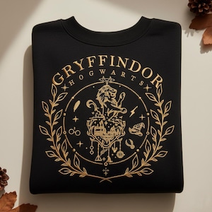 Peut inclure: Sweat-shirt noir avec écusson Gryffondor brodé en or. L'écusson représente un lion, un château et d'autres symboles dans un cercle, entouré d'une couronne de laurier. Le mot "GRYFFINDOR" est inscrit en arc de cercle, avec "HOGWARTS" en dessous.