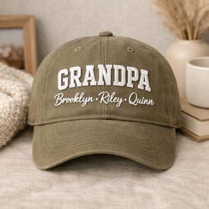 May include: Olive green baseball cap with the word "GRANDPA" in white block letters. Below the word "GRANDPA" are the names "Brooklyn, Riley, Quinn" in white script. The cap is made of a soft fabric.