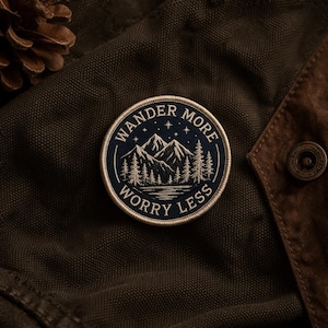 May include: A circular patch with the words "Wander More Worry Less" encircling a mountain and forest scene. The patch is navy blue with white and gold accents, and is set against a dark green fabric.