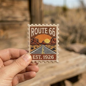 May include: Embroidered patch with a Route 66 design. The patch shows a road leading to a sunset, with the text "ROUTE 66" and "EST. 1926". The patch is held in a hand.