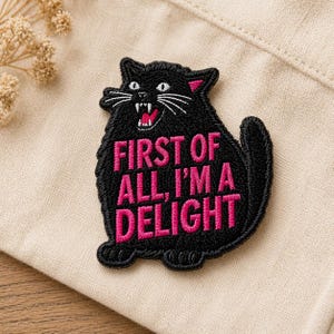 May include: Black cat patch with the text "FIRST OF ALL I'M A DELIGHT" in pink. The cat has white whiskers, pink ears, and an open mouth showing teeth. The patch is on a beige fabric.