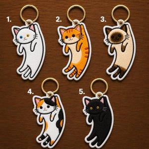 May include: Embroidered cat keychains in various colors: white with blue and yellow eyes, orange tabby, Siamese, calico, and black. Each keychain features a gold-colored ring. The cat designs are outlined in black.