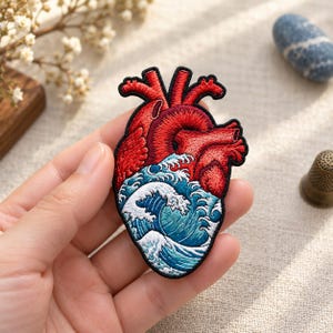 May include: An embroidered patch featuring an anatomical heart design. The upper portion of the heart is rendered in vibrant red thread, while the lower section depicts a stylized blue and white ocean wave pattern with a black stitched border.