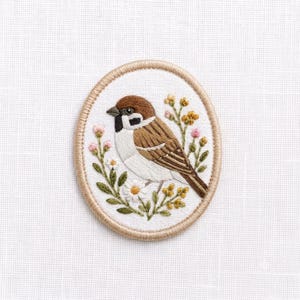 May include: An oval embroidered patch featuring a brown and white sparrow perched among small yellow and pink flowers with green leaves. The design is stitched on a white fabric background and finished with a tan stitched border edge.