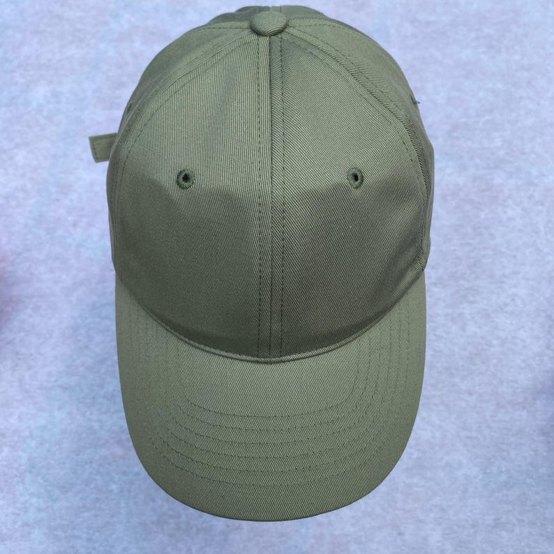 May include: Olive green baseball cap with a curved brim. The cap features a button on top and two small ventilation holes. A fabric strap is visible on the back for adjustment. The cap is made of a durable, solid-colored fabric.