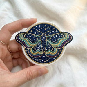 May include: Embroidered patch featuring a celestial moth design. The patch has a navy blue background with gold, teal, and purple accents. The moth is surrounded by stars and a crescent moon. The patch is round with a white border.